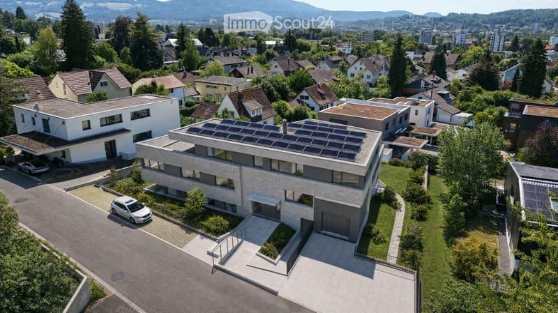 Aerial view of a modern, multi-story house with solar panels on the roof, surrounded by a well-maintained garden and driveway with parked cars. The house features large windows and a contemporary design, situated in a suburban neighborhood with other hous