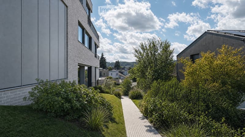 Modern residential complex with a landscaped garden area, featuring neatly trimmed bushes, a paved pathway, and contemporary buildings with large windows and clean lines.