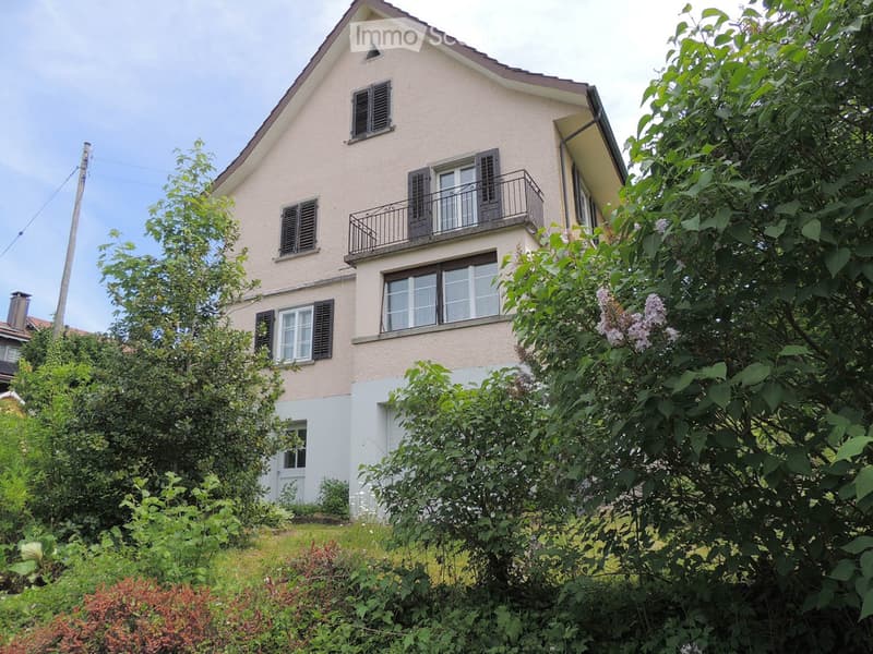 Two-story house with beige siding, multiple windows with shutters, and a small balcony on the upper floor. The house is surrounded by lush greenery and trees.