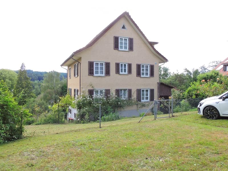 Two-story house with beige siding, multiple windows with shutters, and a triangular roof. The house is surrounded by a fenced yard with grass and trees, and a white car is parked nearby.