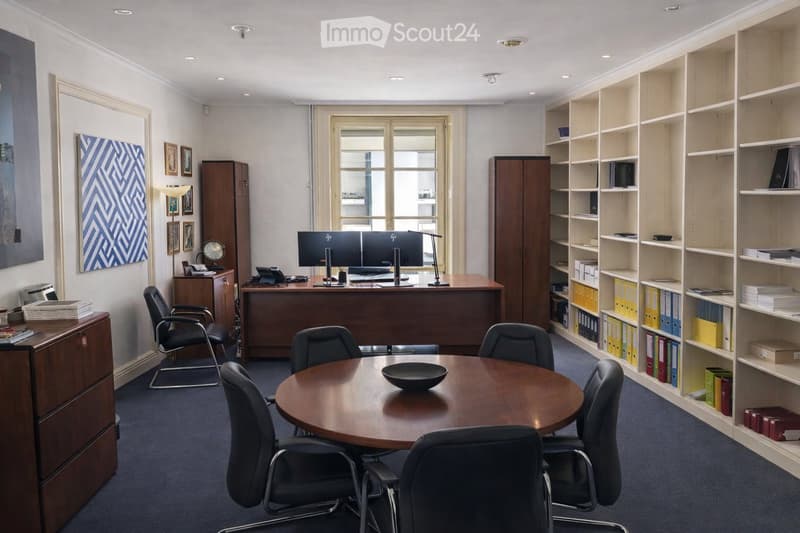 Office space with a large wooden desk, two computer monitors, a round meeting table with chairs, shelves filled with binders and books, filing cabinets, and artwork on the walls.