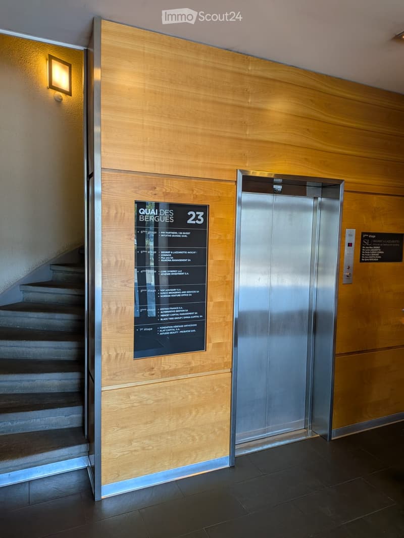 Modern hallway with wooden panel walls, elevator, staircase, and illuminated signage displaying floor numbers and tenant names.