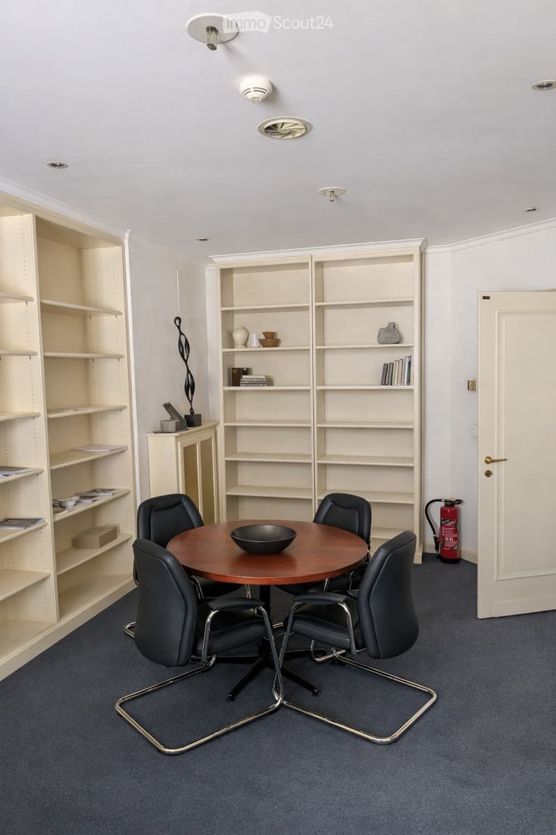 A furnished room with a round wooden table surrounded by black chairs, shelves with books and decorative items, and a fire extinguisher near the door.
