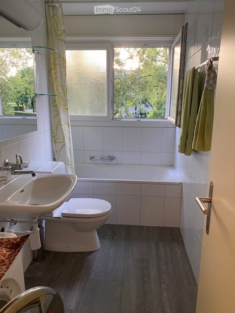 Bathroom with a sink, toilet, bathtub, and a window with a curtain. The room features white tiles and a dark wooden floor.