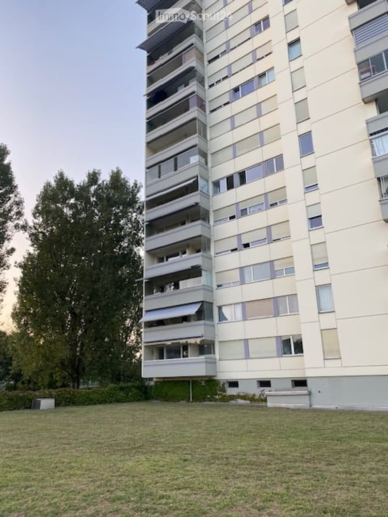 Modern multi-story apartment building with balconies and large windows, surrounded by a grassy area and trees.
