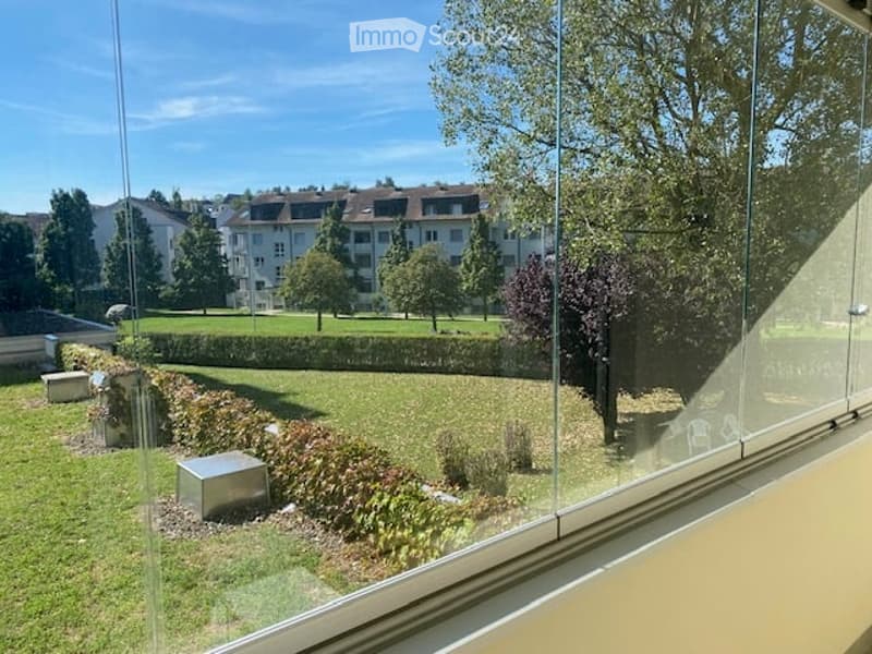 View from a room showing a well-maintained garden with grass, trees, shrubs, and a low hedge. The background includes a residential area with apartment buildings and parked cars.