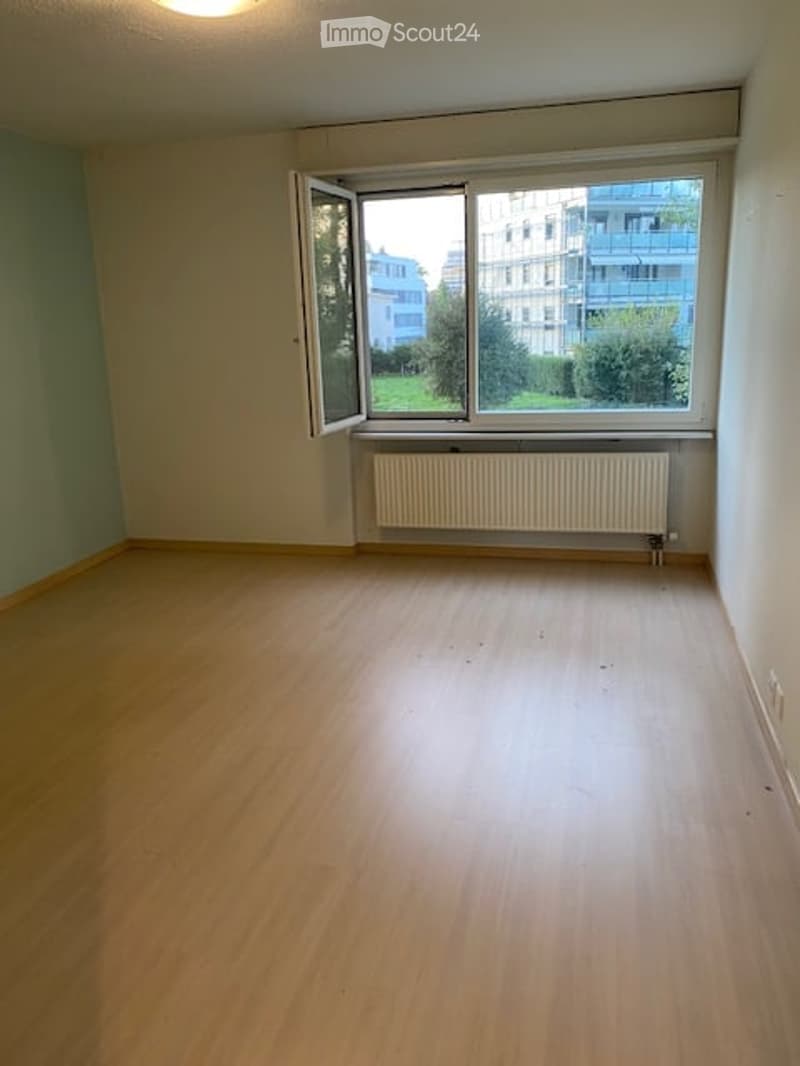 Empty room with wooden flooring, white walls, large window with open shutters, radiator below the window, and a view of greenery and buildings outside.