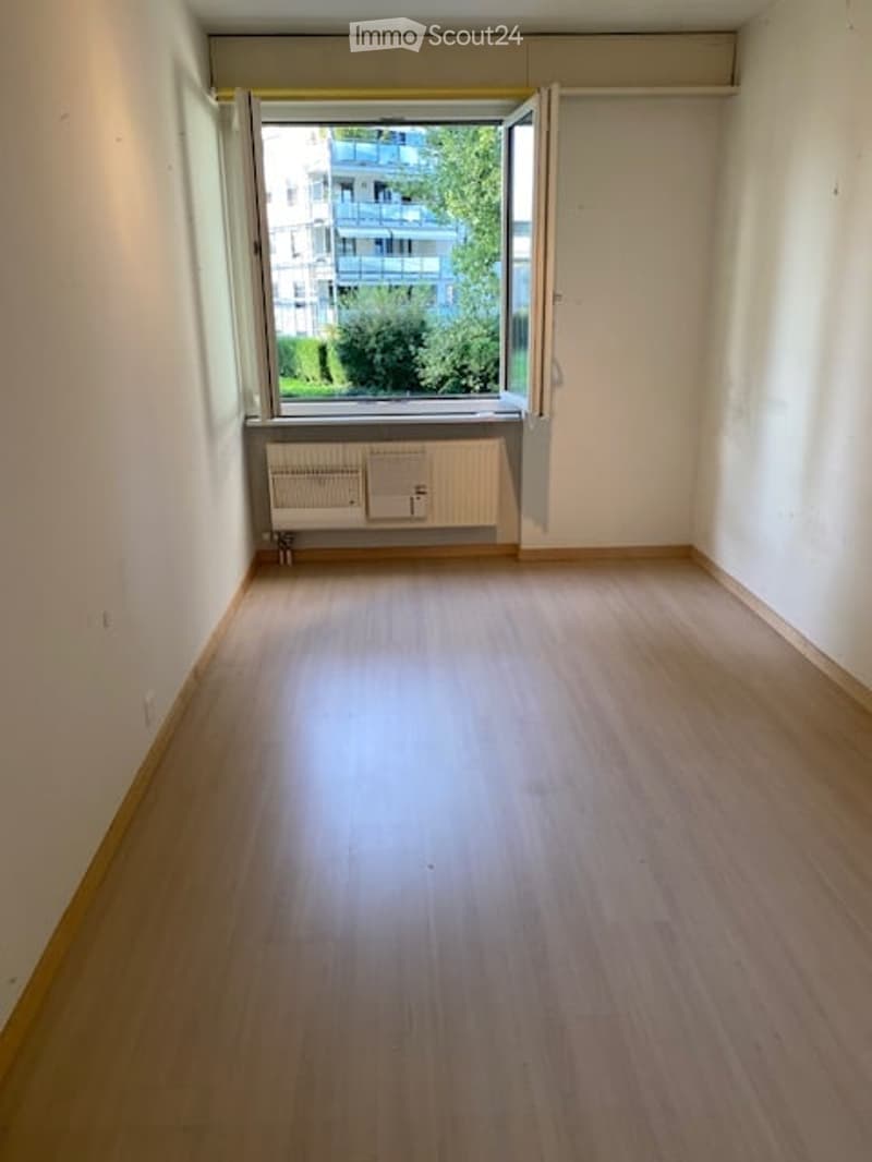 Empty room with wooden flooring, white walls, and a large window with a view of a building and greenery outside. Radiator under the window.