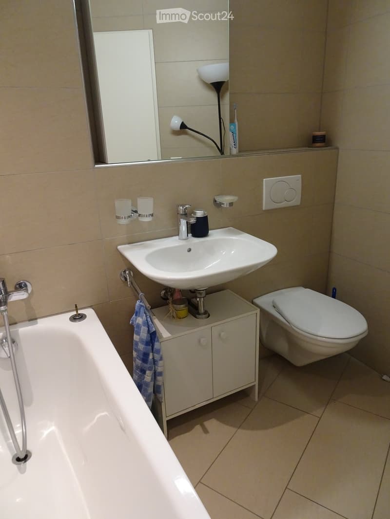 Modern bathroom with a white bathtub, sink, toilet, and a small cabinet. Features a mirror, light fixture, and a towel hanging on the side.