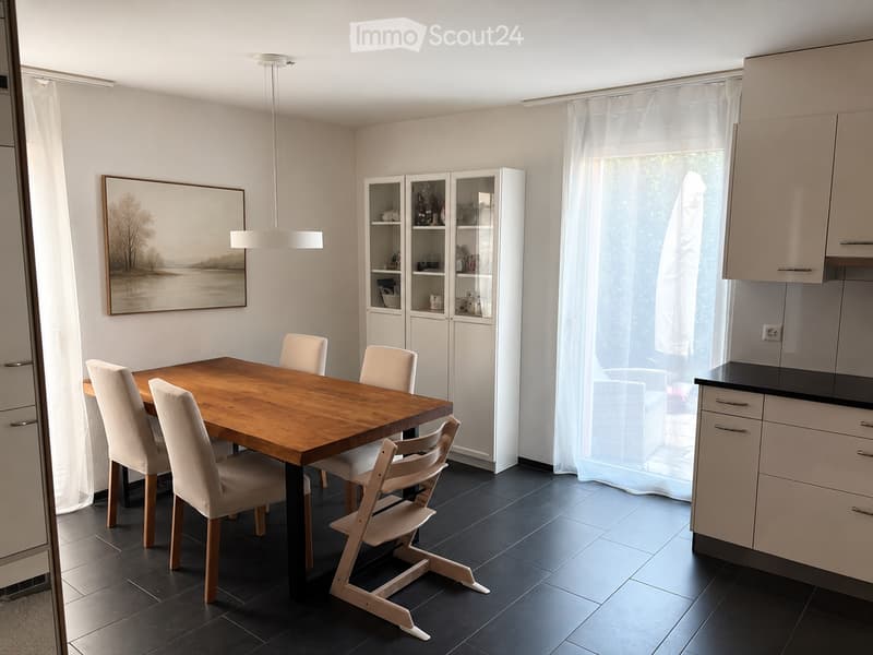 Dining area with a wooden table, six chairs, white cabinets, and a picture on the wall. The room has large windows with white curtains and a modern kitchen area visible in the background.