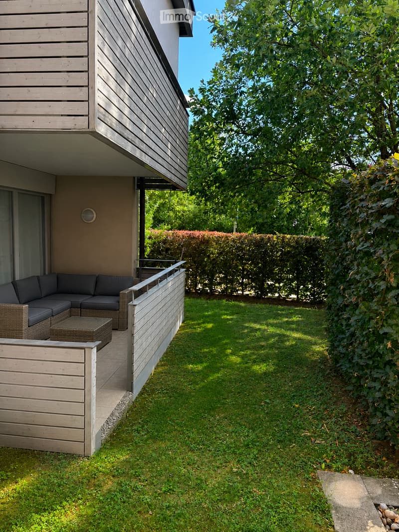 Outdoor patio area with a black sectional sofa, a wicker coffee table, and a wooden railing, surrounded by a grassy lawn and trees.