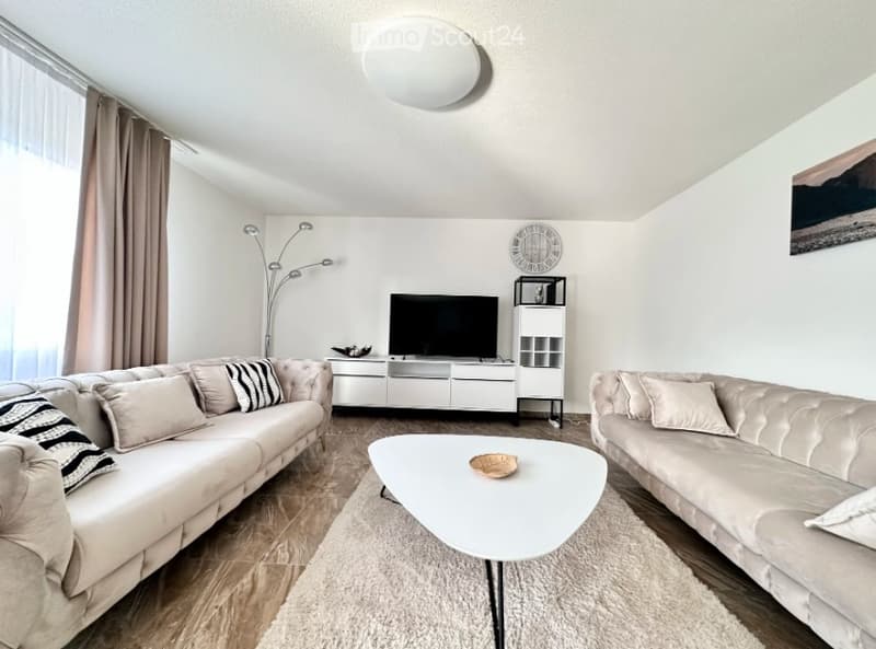 Modern living room with light-colored sofas, a white coffee table, a TV stand, a floor lamp, and a clock on the wall.