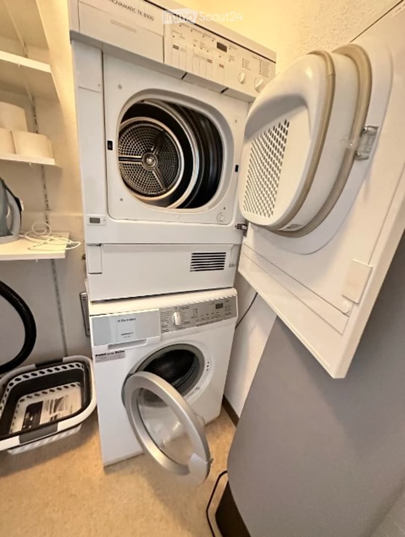 A compact laundry area featuring a stacked washer and dryer unit with open doors, a small shelf holding cleaning supplies, and a laundry basket on the floor.