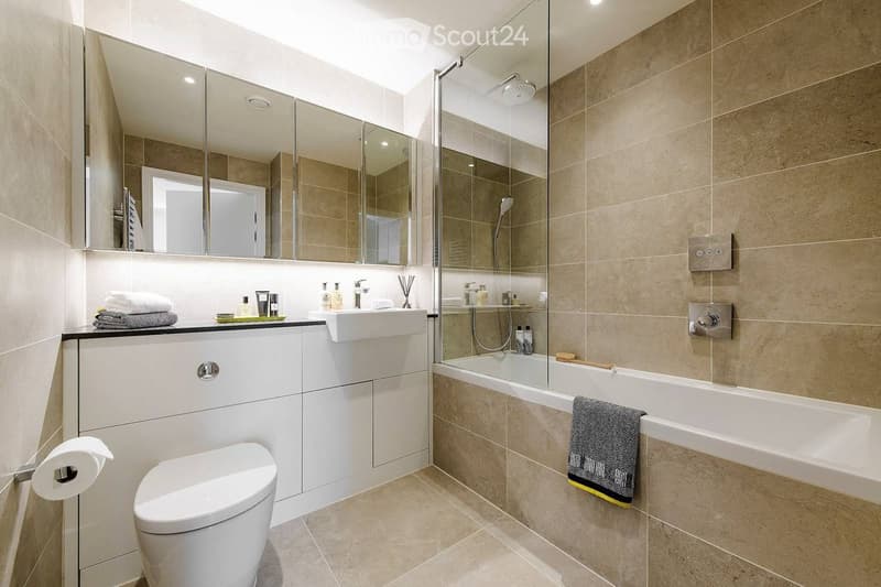 Modern bathroom with beige tiles, a white sink, toilet, bathtub, and a large mirror above the sink. Towels and toiletries are neatly arranged on the counter.