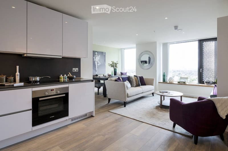 Modern open-plan living and dining area with a sofa, coffee table, dining table, and kitchen area featuring cabinets and an oven. Large windows provide natural light, and decorative items such as vases and a mirror are present.