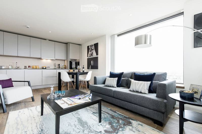 Modern living room with grey sectional sofa, coffee table, white dining chairs, and a sleek kitchen area with cabinets and a sink.