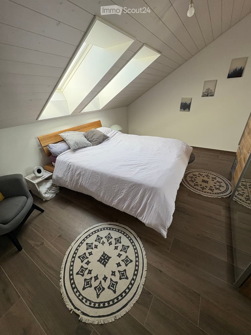 Cozy attic bedroom with wooden slanted ceilings, skylights, a large bed with pillows and blankets, a nightstand with a lamp, a round patterned rug, and a grey armchair.