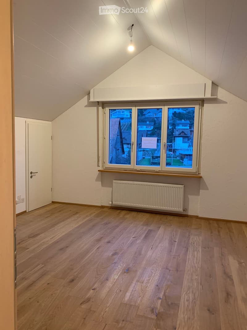 Empty attic room with wooden flooring, white walls, large window, radiator, and a door leading to another area.