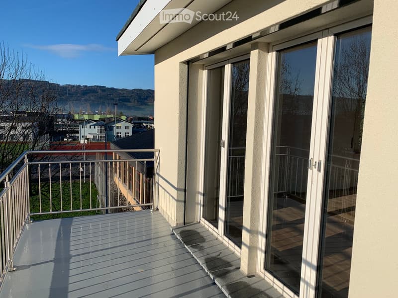 Balcony with wooden decking, metal railing, large glass doors leading inside, scenic view of landscape and buildings.