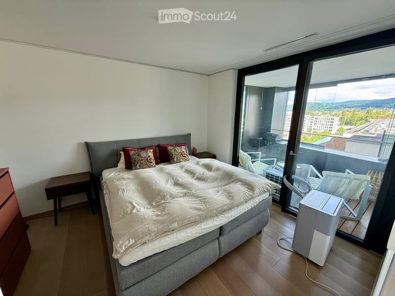Modern bedroom with a large bed, nightstand, and a sliding glass door leading to a balcony with outdoor seating.