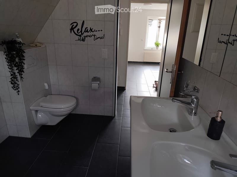 Modern bathroom with a toilet, sink, and faucet. The walls are tiled, and there is a decorative plant on the cabinet. The floor is dark tiled.