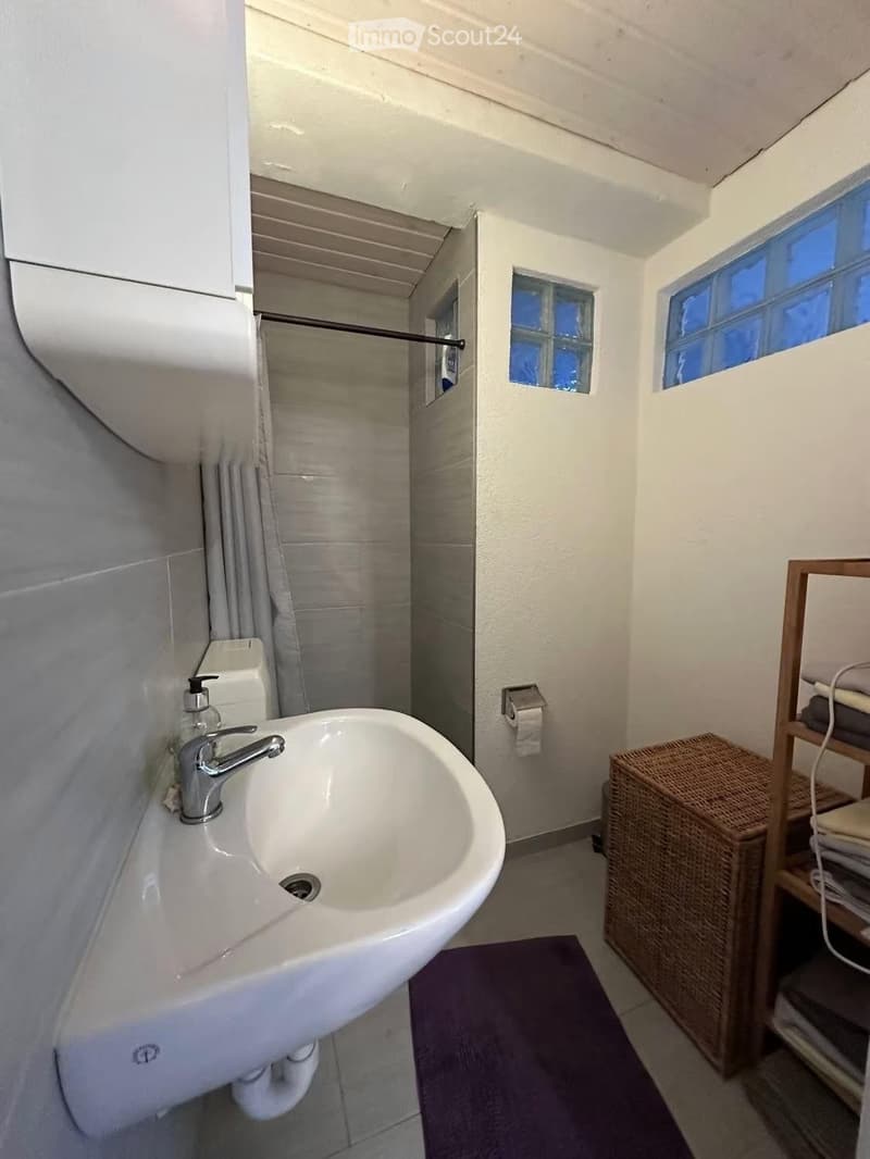 A small bathroom with a white sink, chrome faucet, toilet, and a wicker basket on a wooden shelf. The room has white walls, a purple bath mat, and small windows with frosted glass.