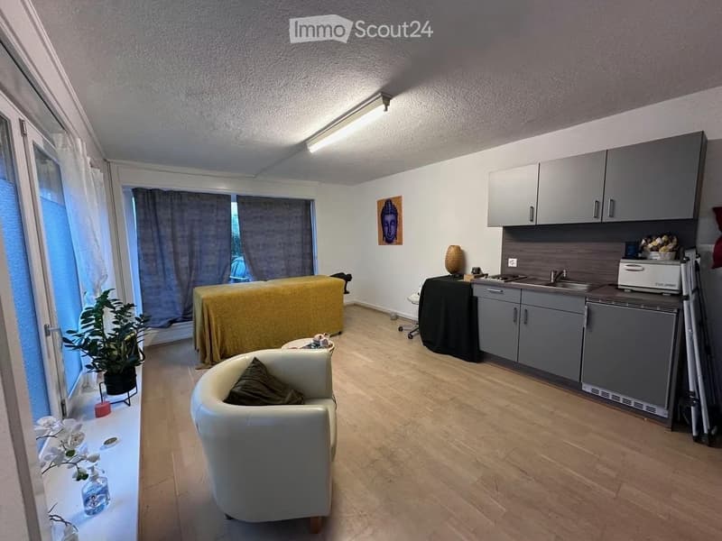 A modern, well-lit room with a sofa, a small table, a kitchen area with cabinets, sink, and fridge, and a massage table covered with a yellow sheet.