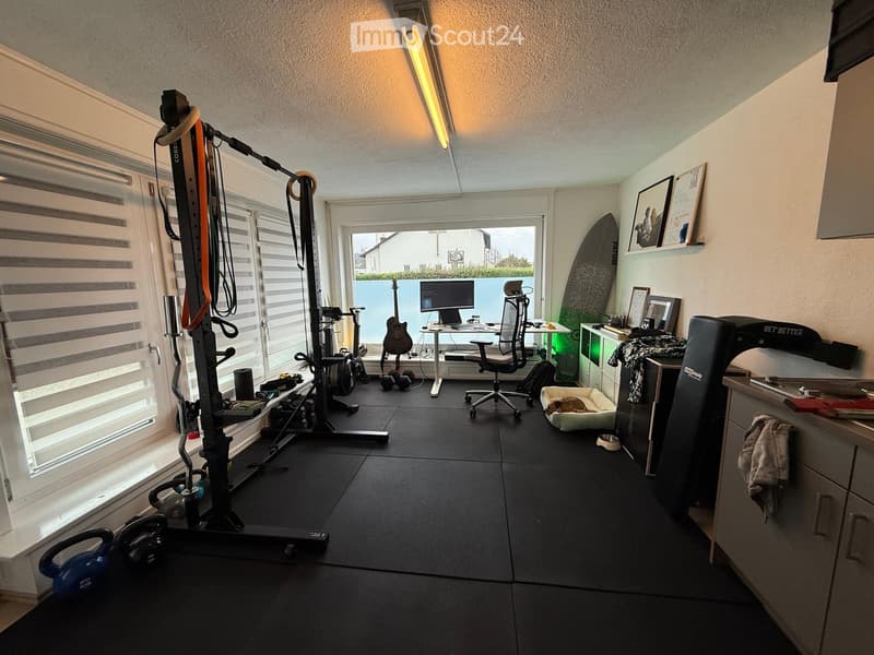 Well-lit room with exercise bike, treadmill, guitar, computer, chair, dog bed, framed pictures, surfboard, kettlebell, and headphones.