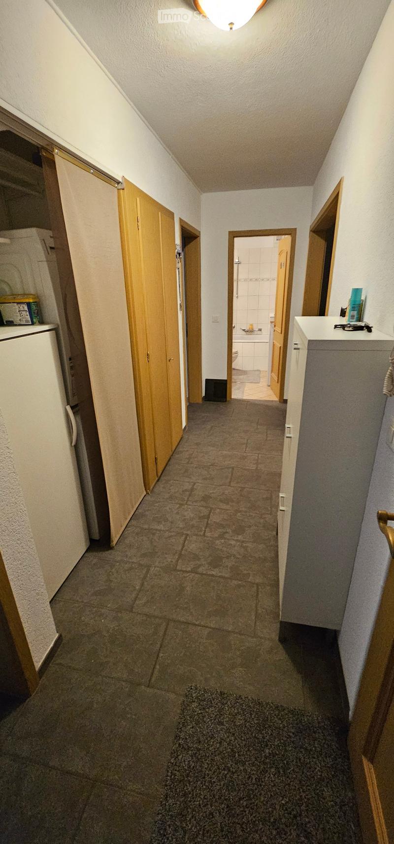 A narrow hallway with tiled flooring, wooden doors, and visible rooms including a bathroom and a kitchen area.