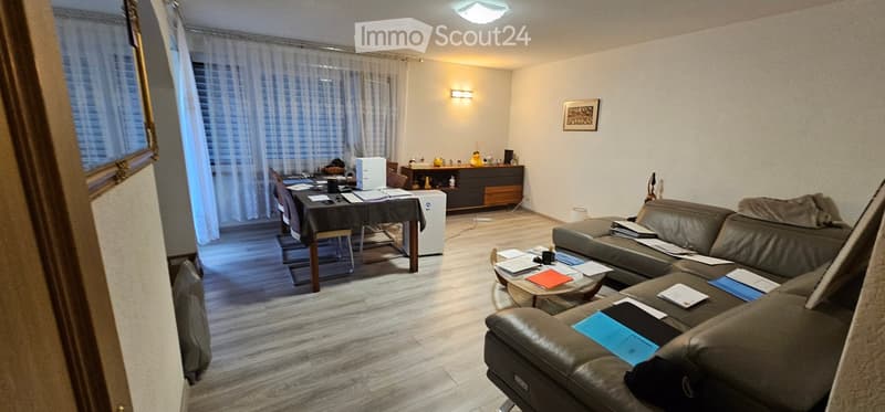 Living room with a grey leather sofa, coffee table, dining area with a table and chairs, a small refrigerator, and a cabinet with decorative items.