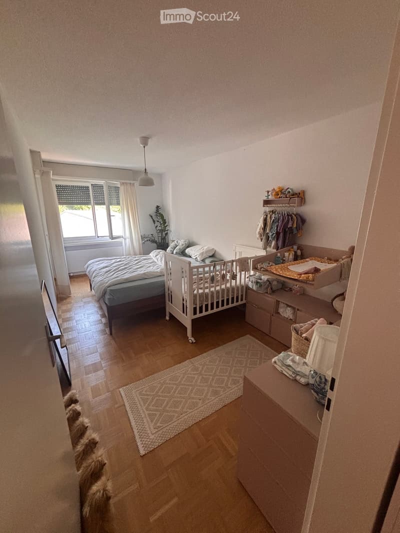 Brightly lit bedroom with wooden flooring, a double bed, a crib, a changing table, a wardrobe, and a small rug.