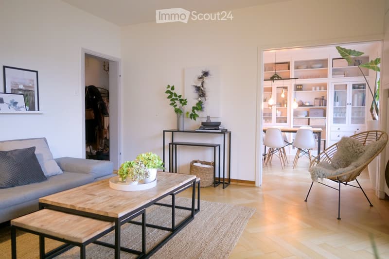 Modern living room with light wooden flooring, grey sofa, wooden coffee table, side table, dining area with white chairs and table, and decorative plants.