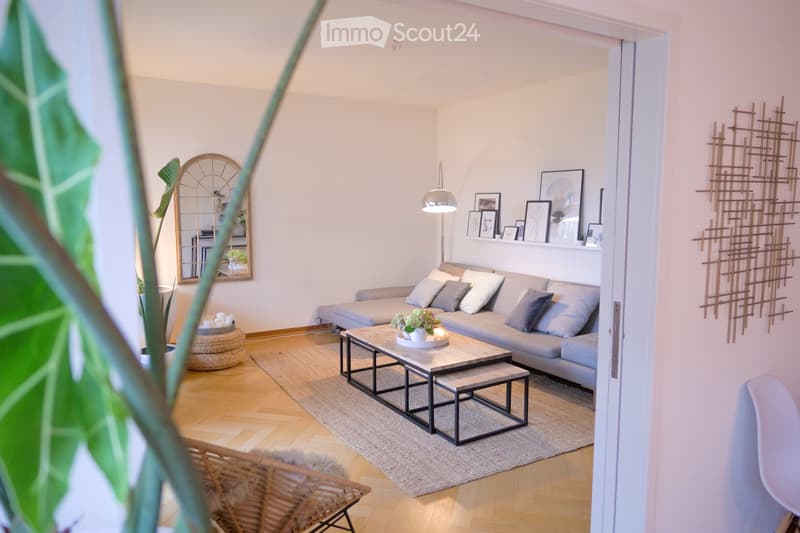 Modern living room with light-colored walls, wooden flooring, and a grey sectional sofa. Features a coffee table with a vase and candle, a wicker chair, a mirror, and wall art.