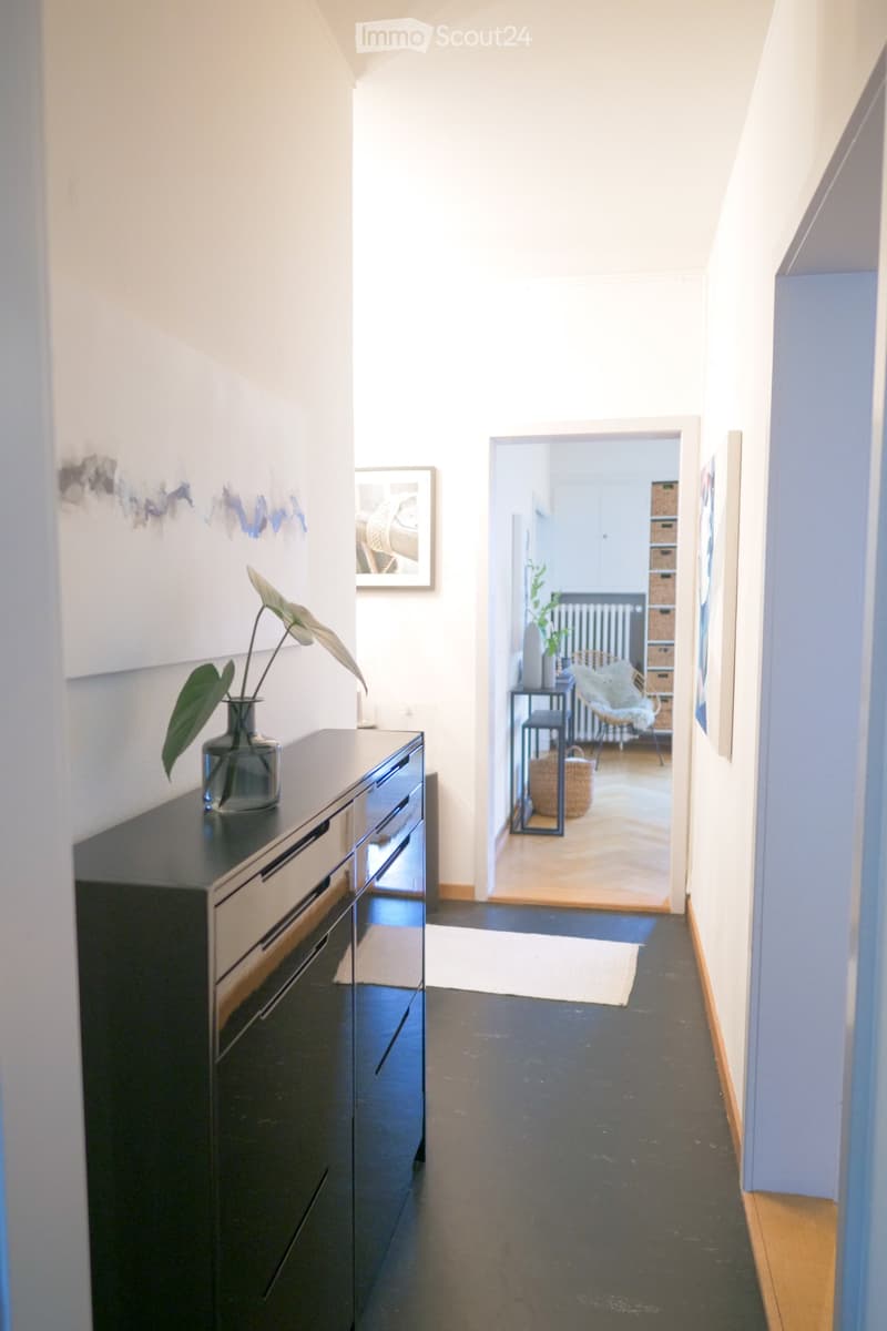 A modern hallway with dark flooring, white walls, and a sleek black console table with a vase of green leaves. An open doorway leads to a room with a wicker chair, a small table, and a radiator.
