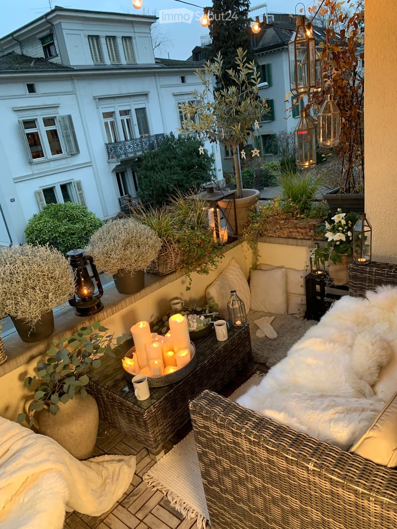 Cozy balcony decorated with wicker furniture, candles, and potted plants, featuring a white fur throw and lanterns.