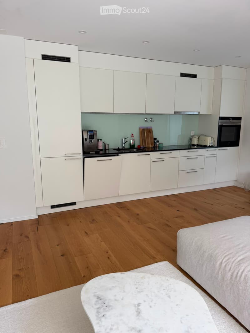 Open-plan living and sleeping area with a modern kitchen featuring white cabinets, stainless steel appliances, and a marble coffee table in front of a bed.