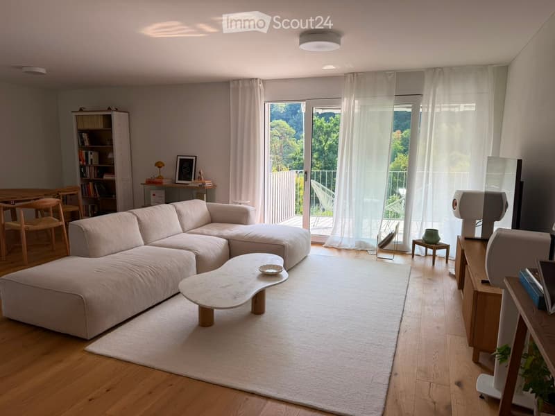 Modern living room with a large sectional sofa, coffee table, dining area, bookshelf, and large windows leading to a balcony.