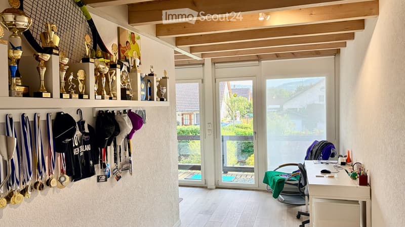 A well-lit room with a wooden ceiling, trophies and medals on a shelf, sports ribbons and hats hanging on the wall, a desk with a chair, and a large window with a view of the garden.