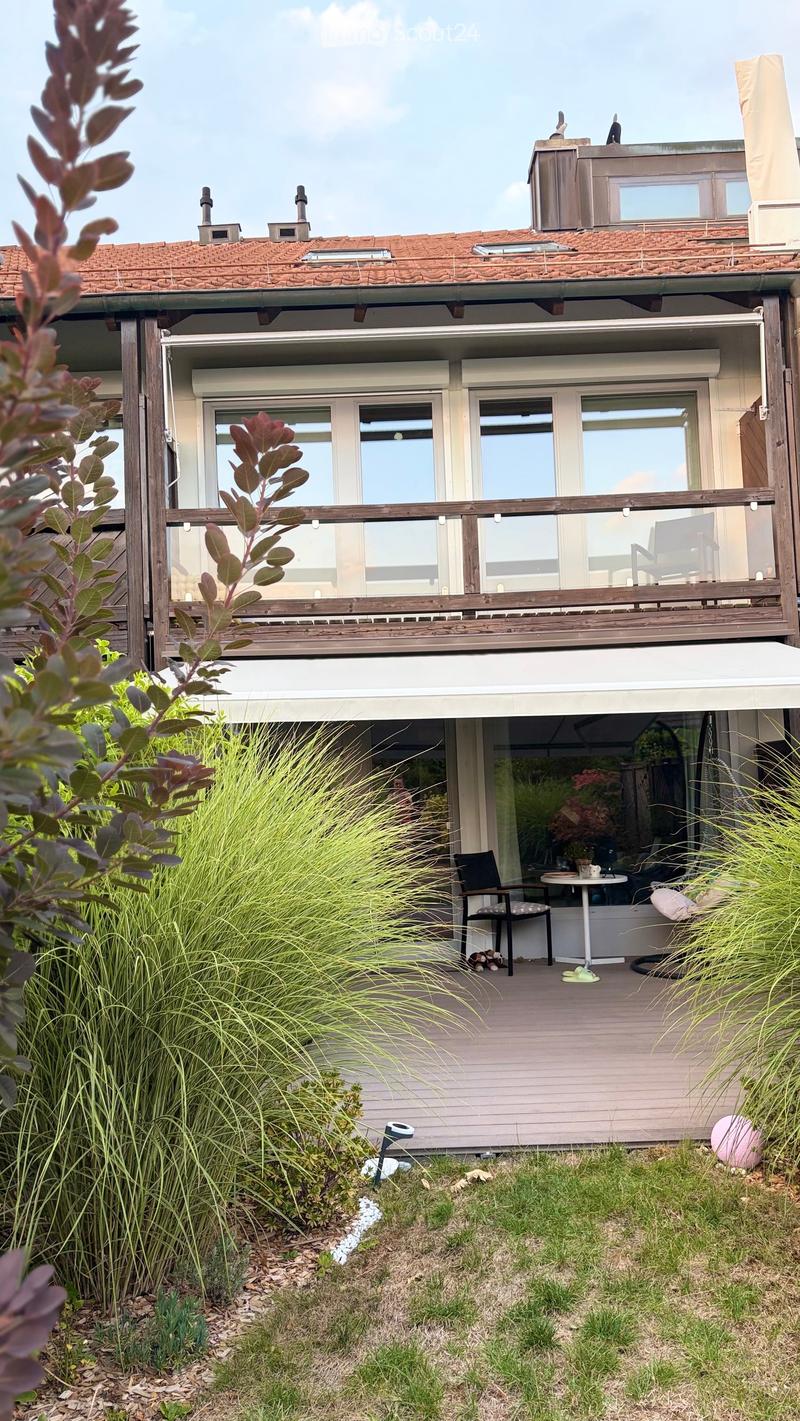 A modern two-story house with a wooden balcony and a well-maintained garden area featuring tall grasses, shrubs, and a small seating area with a table and chairs.