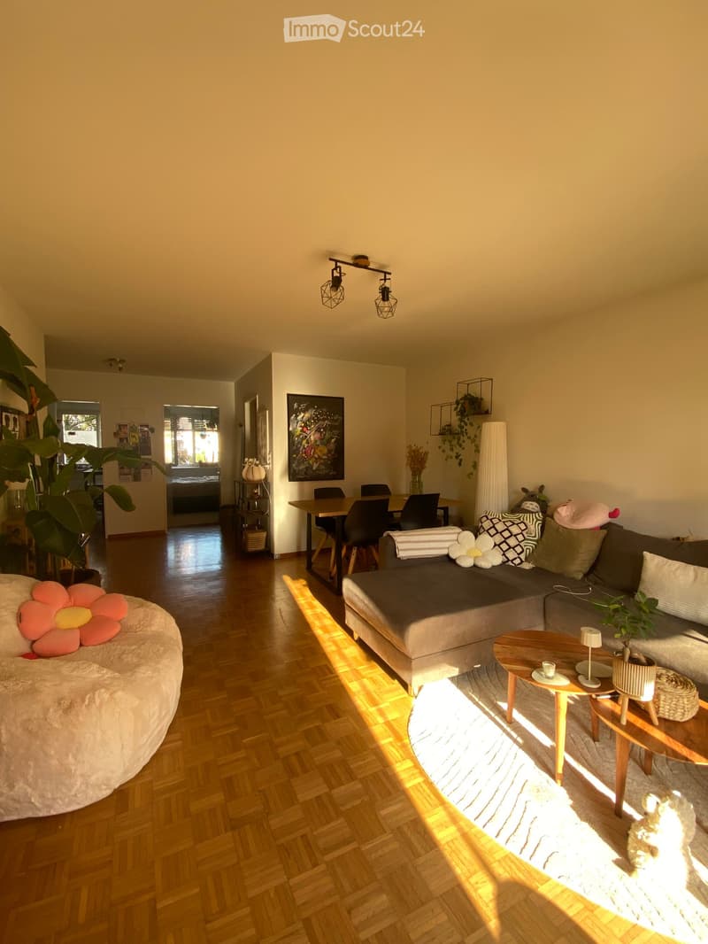 Bright living room with wooden flooring, a grey sectional sofa, a round coffee table, a dining table with chairs, and decorative plants.