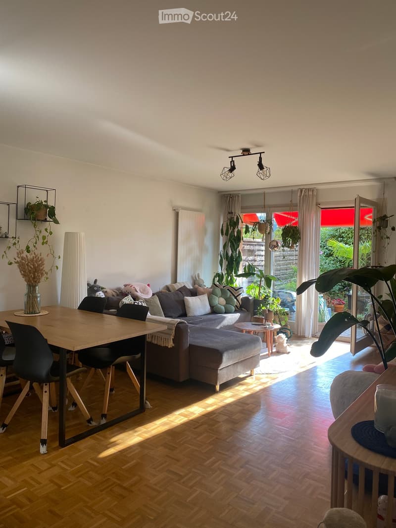 Bright living room with wooden flooring, large window with sheer curtains, and a sliding glass door leading to a garden. Features a grey sectional sofa with multiple cushions, a wooden coffee table, a dining table with chairs, and several potted plants.