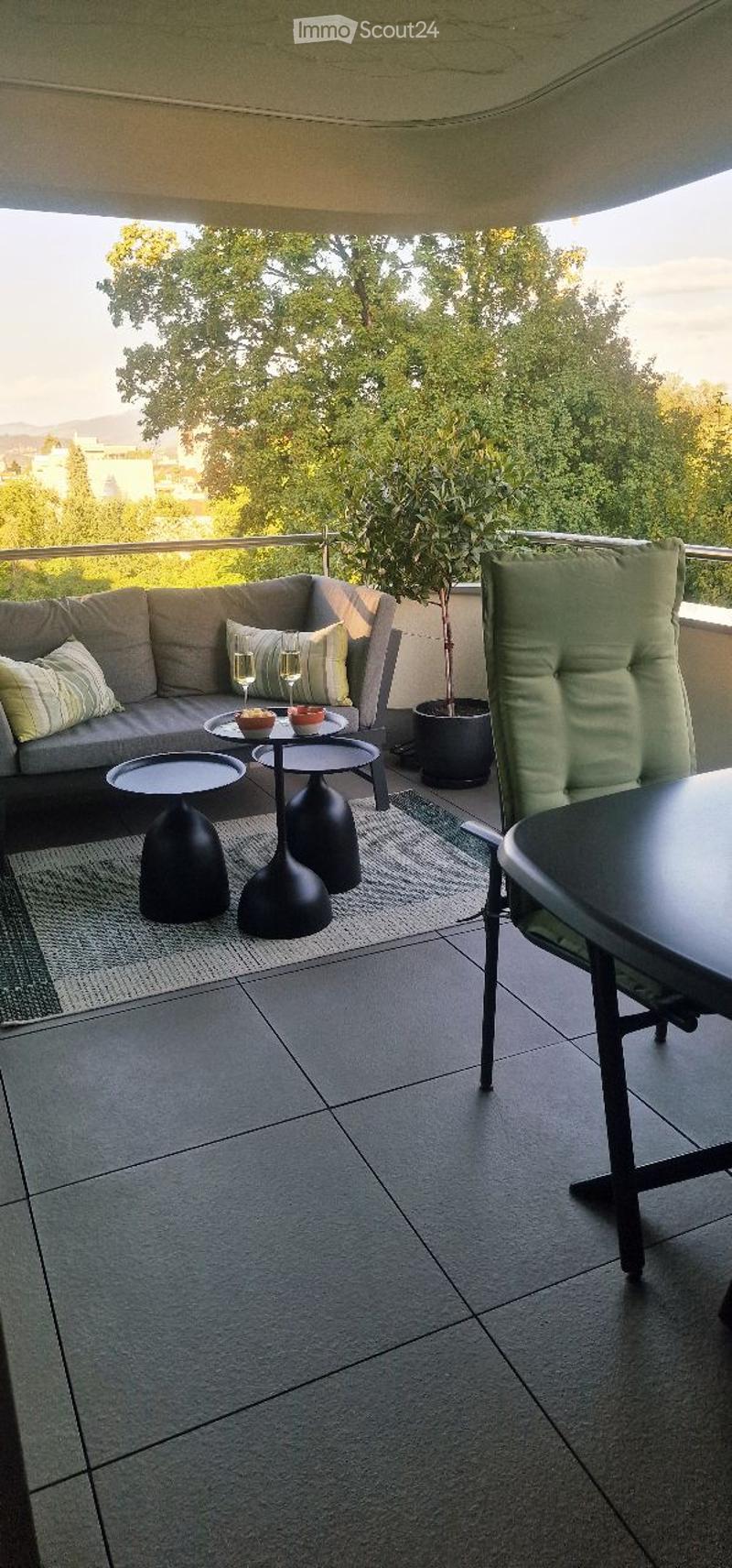 Balcony with grey sofa, black tables, and a potted plant, overlooking a green view.