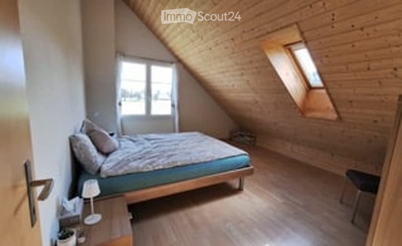 Cozy attic bedroom with wooden floors, slanted ceilings, large windows, a bed with pillows and blankets, a nightstand with a lamp, and a small chair.