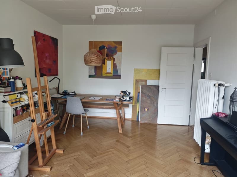 A well-lit room with wooden flooring, a sofa, a chair, a table, a lamp, a radiator, and several paintings on the walls.