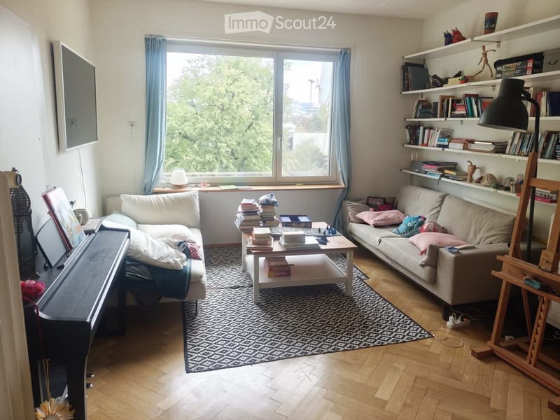 Living room with wooden flooring, large window with blue curtains, two sofas, coffee table with books, bookshelf, and a piano.