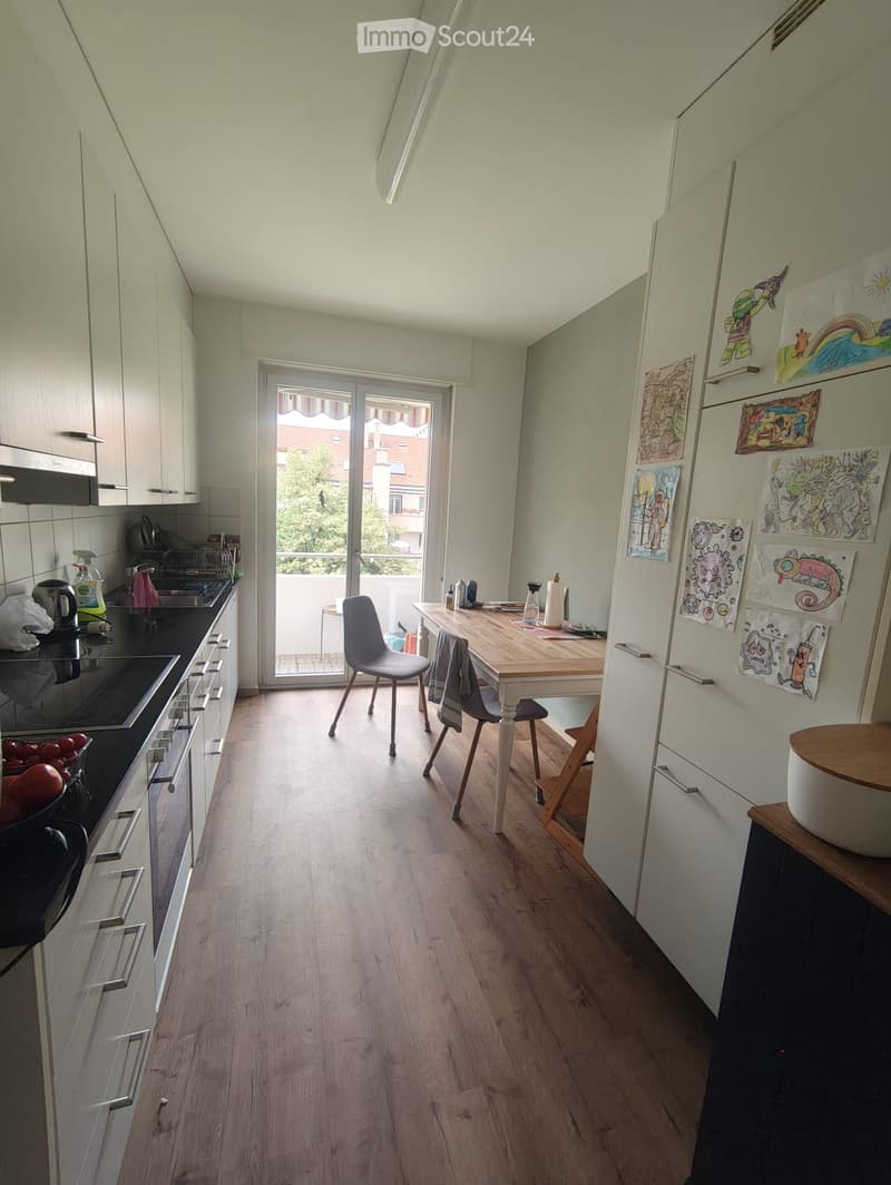 Modern kitchen with light wooden flooring, white cabinets, and a dining area with a table and chairs. Features a balcony door leading outside.
