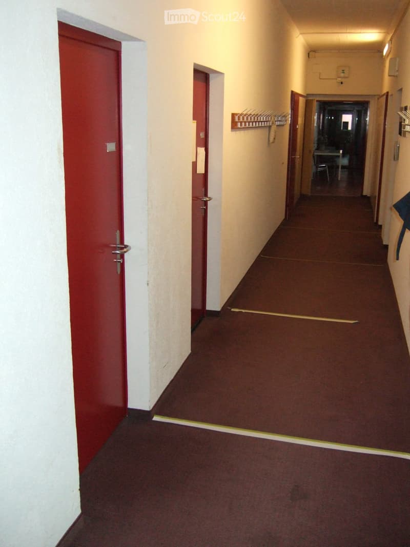 A narrow hallway with red carpeting, white walls, and several doors, including a red door on the left and a coat rack on the right.