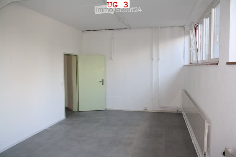 Empty room with white walls, grey concrete floor, and a light green door leading to another space. Windows on the right side allow natural light to enter.