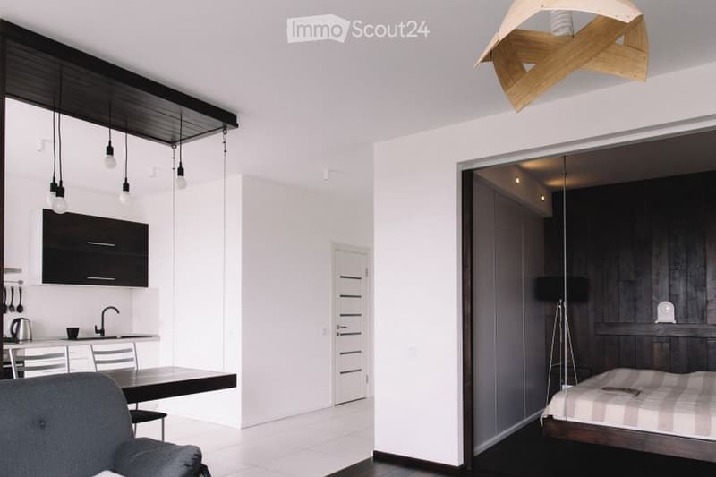Modern open-plan living space with a kitchen area, dining table, and a separate bedroom area with a bed and lamp.