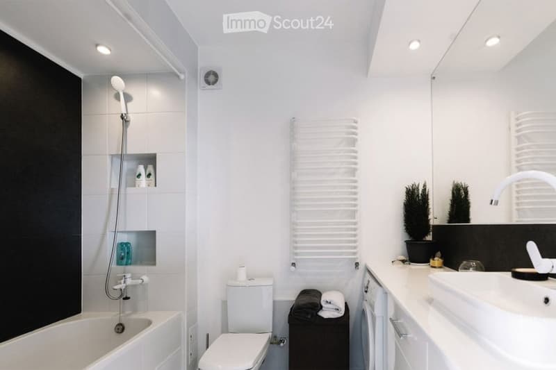 Modern bathroom with white tiles, bathtub, toilet, sink, towel rack, and small potted plants on the counter.